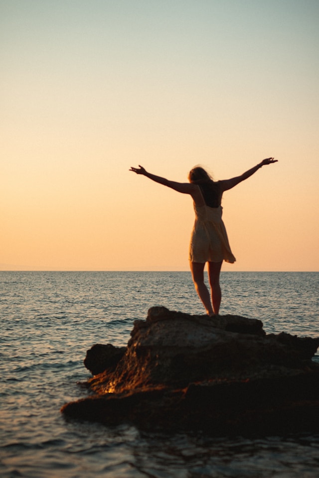 Person mit ausgebreiteten Armen auf einem Felsen am Meer bei Sonnenuntergang